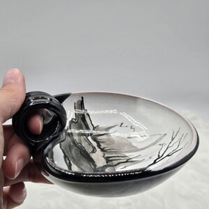 May include: A ceramic bowl with a black handle and rim. The interior features a landscape design in shades of gray and black. The bowl is held by a hand, showcasing its unique shape and artistic details.