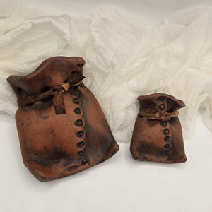 May include: Two brown ceramic pouches with a rustic appearance. Each pouch is tied with a ribbon and features a row of decorative buttons down the front. The pouches are of different sizes, suggesting a set.