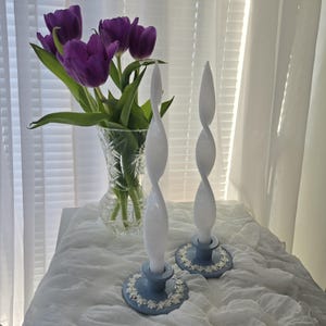 May include: Two white, twisted taper candles on light blue candle holders with a floral design. A bouquet of purple tulips in a crystal vase is in the background. The scene is set on a white cloth near a window.