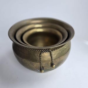 May include: Set of three antique brass bowls, nested inside each other. The bowls have a rounded shape with a wide rim and a decorative rope detail around the top. The brass has a warm, aged patina.