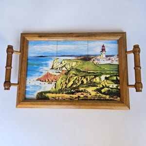 Può includere: Vassoio di legno con mosaico di piastrelle raffigurante un paesaggio costiero. Il mosaico mostra un faro, scogliere e il mare. Il vassoio è rettangolare con manici in legno.