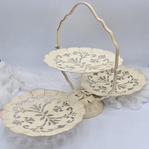 May include: A three-tiered serving tray with a handle. The tray is off-white with a floral cut-out design on each tier. The metal frame has a distressed finish, giving it a vintage look. Ideal for displaying desserts or appetizers.