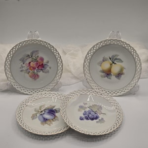 May include: Four white porcelain dessert plates with a delicate, openwork border. Each plate features a different fruit design: strawberries, apples, plums, and grapes. The fruit illustrations are in soft, realistic colors, with gold trim.
