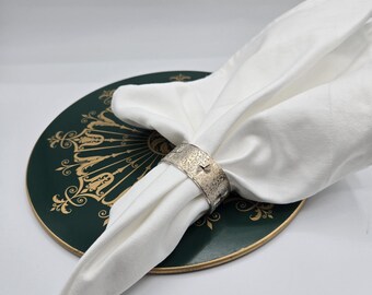 May include: A white linen napkin is held by a silver napkin ring with a textured finish. The napkin ring sits on a dark green plate with gold decorative patterns and a gold rim.