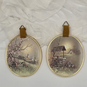 May include: Two oval wall art pieces with gold frames and landscape paintings. One depicts a village scene with a church, the other a well. Each has a brown leather strap for hanging.
