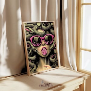 May include: A framed art print featuring a stylized illustration of Medusa wearing pink sunglasses and enjoying a lollipop. The artwork showcases a woman with snakes for hair, a playful take on the mythological figure. The print is displayed on a wooden surface.