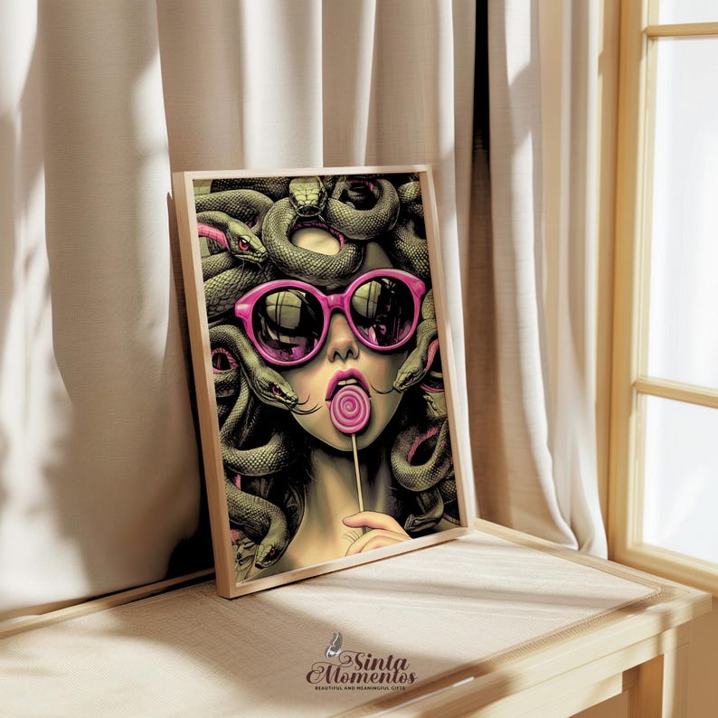 May include: A framed art print featuring a stylized illustration of Medusa wearing pink sunglasses and enjoying a lollipop. The artwork showcases a woman with snakes for hair, a playful take on the mythological figure. The print is displayed on a wooden surface.