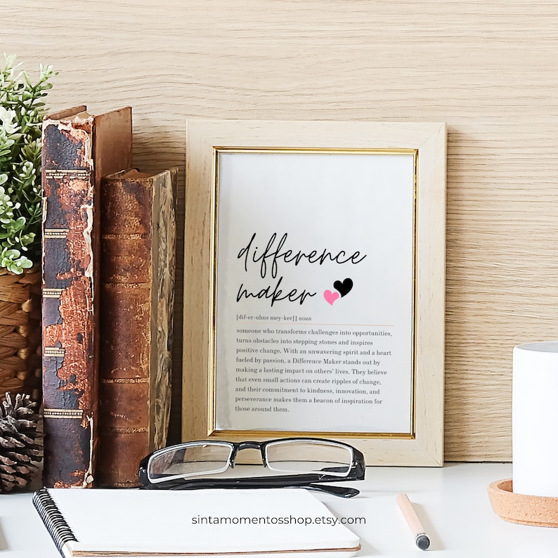 May include: A framed print with a white background and black text that reads "difference maker" with two pink hearts. The text below defines the term "difference maker" as someone who transforms challenges into opportunities, turns obstacles into stepping stones, and inspires positive change. The print is on a wooden table with a notebook and glasses.