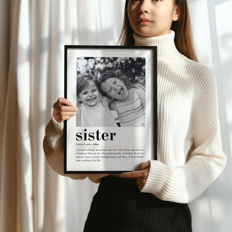 May include: Black and white photograph of two sisters smiling and hugging. The photo is framed with a black frame and has a definition of the word "sister" below the photo.