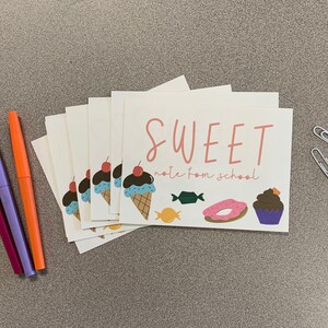 May include: A stack of cream-colored cards with the words "SWEET note from school" in pink and brown. The cards feature illustrations of ice cream cones, candy, donuts, and cupcakes. Three pens and two paper clips are also visible.