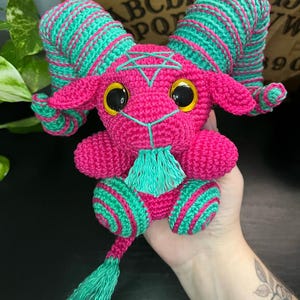 May include: A pink crocheted goat with green and pink striped horns, a green beard, and a green tail. The goat has large black eyes and a pentagram on its forehead.