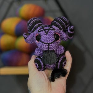 Może przedstawiać: Fioletowa pluszowa zabawka amigurumi z czarnymi rogami, pentagramem na klatce piersiowej i czarnymi oczami. Zabawka ma czarny ogon i siedzi w dłoni osoby.