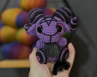 Könnte beinhalten: Lilafarbenes Amigurumi-Spielzeug aus Häkelarbeit mit schwarzen Hörnern, einem Pentagramm auf der Brust und schwarzen Augen. Das Spielzeug hat einen schwarzen Schwanz und sitzt in der Hand einer Person.