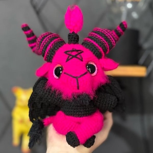 Op de afbeelding: Een roze gehaakt pluche speelgoed met zwarte hoorns, een zwarte baard en een zwart pentagram op zijn voorhoofd. Het speelgoed heeft grote zwarte ogen en een klein roze hoorn op zijn hoofd.