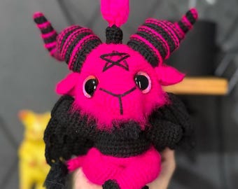 May include: A pink crocheted plush toy with black horns, a black beard, and a black pentagram on its forehead. The toy has large black eyes and a small pink horn on top of its head.
