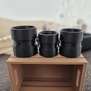 May include: Three black cylindrical objects with a stepped design, each with a small hole, are displayed on a small brown wooden crate. The objects vary in size, with the largest on the left and the smallest in the center.