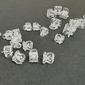 May include: A collection of clear plastic hair clips, also known as mini claw clips, scattered on a gray surface. The clips are small and have a variety of shapes and sizes.