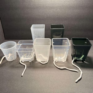 May include: A collection of clear and dark green plastic plant pots with attached white wicks. The pots vary in shape, including round and square designs, and are arranged on a dark surface. These pots are designed for self-watering.