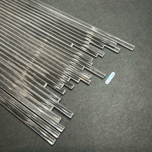 May include: A collection of clear, rectangular acrylic rods, approximately 0.12 inches thick, arranged on a dark gray surface. The rods are transparent and appear to be cut to various lengths. The image is well-lit, highlighting the clarity of the material.