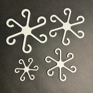May include: Four white plastic snowflake-shaped ornaments of varying sizes. The ornaments have six points each, with a small loop at the end of each point.