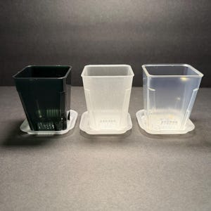 May include: Three square plastic plant pots in varying shades: dark green, frosted white, and clear. Each pot sits on a matching square saucer. The pots are designed with drainage holes and a tapered shape.