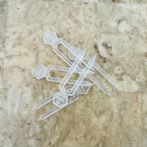 May include: A set of six clear plastic plant markers with a hexagonal shape and a pointed end. The markers have the text "Plant Marker" printed on them.