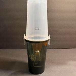 Puede incluir: Una maceta de plástico transparente con una base verde oscuro y una tapa translúcida. La maceta tiene forma cilíndrica con una parte superior ligeramente ensanchada. La tapa tiene pequeños orificios de ventilación. La maceta está diseñada para la propagación de plantas en interiores.