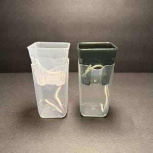May include: Two clear plastic orchid pots with a dark green top and a clear plastic pot. The pots have a square top and a clear plastic insert with a white string. The pots are designed for orchids.