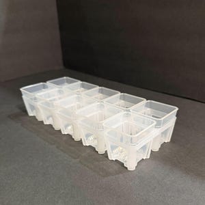May include: A clear plastic seedling tray with twelve individual square cells. Each cell has a raised rim and is supported by four small legs. The tray is designed for starting plants indoors.