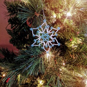 May include: A white crocheted snowflake ornament with blue beads hangs on a green Christmas tree with white lights.