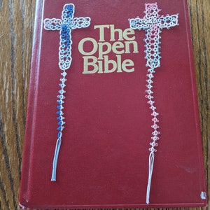 Può includere: Due segnalibri in pizzo bianco a forma di croce, uno blu e uno rosa, su un libro rosso con il titolo "The Open Bible" in lettere dorate.