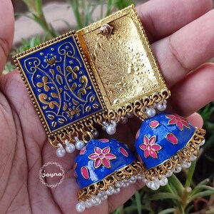 Pode incluir: Um par de brincos azuis e dourados com detalhes florais rosa. Os brincos s&atilde;o feitos de metal e t&ecirc;m um design pendente de v&aacute;rias camadas. Os brincos s&atilde;o decorados com pequenas contas brancas.