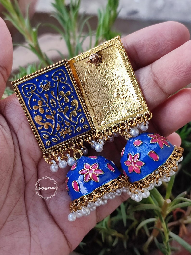 Pode incluir: Um par de brincos azuis e dourados com detalhes florais rosa. Os brincos s&atilde;o feitos de metal e t&ecirc;m um design pendente de v&aacute;rias camadas. Os brincos s&atilde;o decorados com pequenas contas brancas.