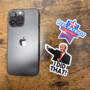 May include: A black iPhone with a white Apple logo and a camera lens on the back. The phone is sitting on a wooden surface with three stickers. One sticker is a red, white and blue star with the text "Peel Patriot Custom Stickers". Another sticker is a photo of a man in a suit with the text "I Did That!" The third sticker is a red, white and blue star with the text "Peel Patriot Custom Stickers".