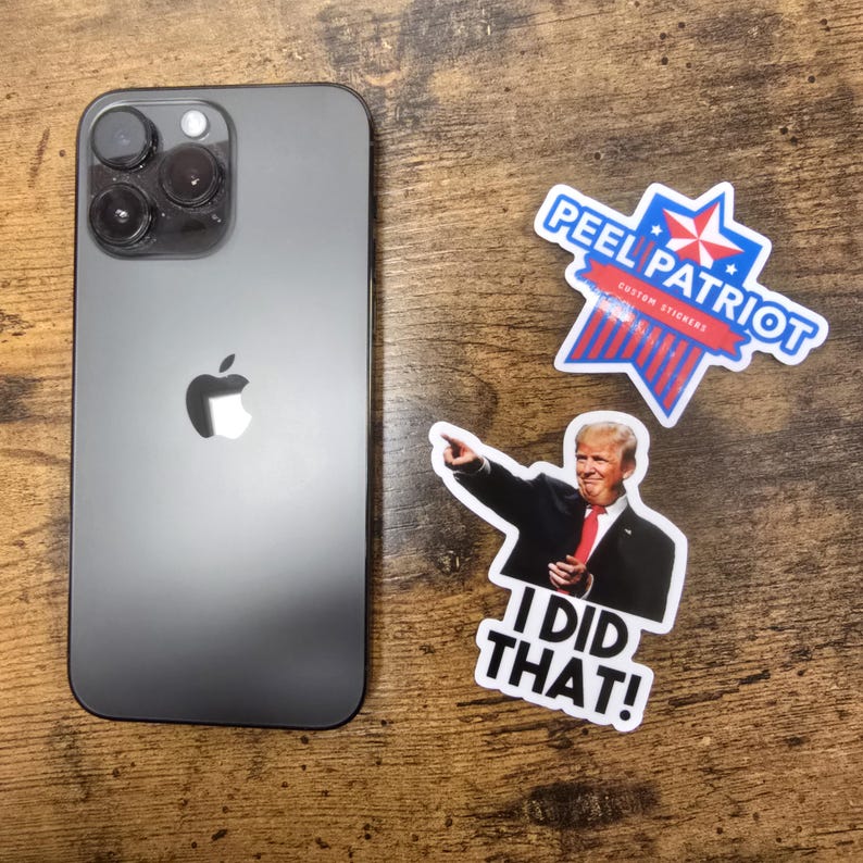 May include: A black iPhone with a white Apple logo and a camera lens on the back. The phone is sitting on a wooden surface with three stickers. One sticker is a red, white and blue star with the text "Peel Patriot Custom Stickers". Another sticker is a photo of a man in a suit with the text "I Did That!" The third sticker is a red, white and blue star with the text "Peel Patriot Custom Stickers".