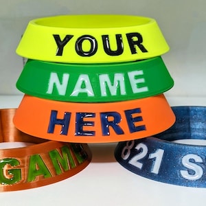 May include: Four 3D printed bowling pin holders. The top holder is bright yellow with black text that says "YOUR NAME HERE". The next holder is green with white text that says "NAME HERE". The third holder is orange with blue text that says "HERE". The bottom holder is blue with white text that says "821 SERIES".