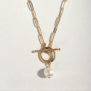 May include: Gold-coloured necklace with a toggle clasp and a dangling white pearl pendant. The chain features elongated oval links. The necklace is displayed against a white background.