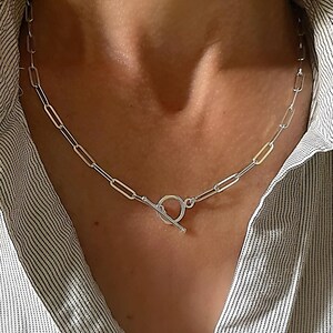 May include: A silver chain necklace with an elongated oval link design. The necklace has a toggle clasp. The necklace is worn against a striped shirt.