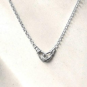 Puede incluir: Un collar plateado con una cadena delicada y un colgante central. El colgante presenta un dise&ntilde;o de dos manos entrelazadas, que simbolizan la unidad o la conexi&oacute;n. El collar se muestra sobre un fondo neutro.
