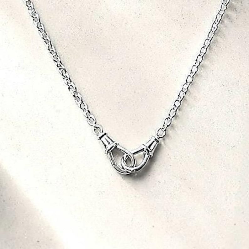 Puede incluir: Un collar plateado con una cadena delicada y un colgante central. El colgante presenta un dise&ntilde;o de dos manos entrelazadas, que simbolizan la unidad o la conexi&oacute;n. El collar se muestra sobre un fondo neutro.