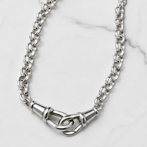 May include: A silver necklace with a chain-link design. The necklace has a distinctive clasp with a looped design. The jewellery is displayed on a white marble surface.