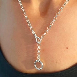 Peut inclure: Un collier argenté composé d'une chaîne à maillons ronds avec un fermoir à bascule central. La chaîne se divise en forme de Y et se termine par un pendentif en forme d'anneau circulaire qui pend verticalement de la structure principale.