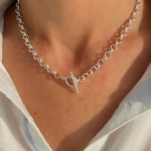May include: A silver-toned chain necklace featuring a series of uniform circular links. The necklace is secured at the front with a toggle clasp, consisting of a horizontal bar that passes through a circular ring, creating a clean and metallic look.