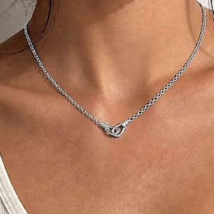 Puede incluir: Un collar plateado con una cadena delicada y un dise&ntilde;o de cierre &uacute;nico. El cierre presenta dos bucles entrelazados, creando una est&eacute;tica moderna y minimalista. El collar se lleva alrededor del cuello.