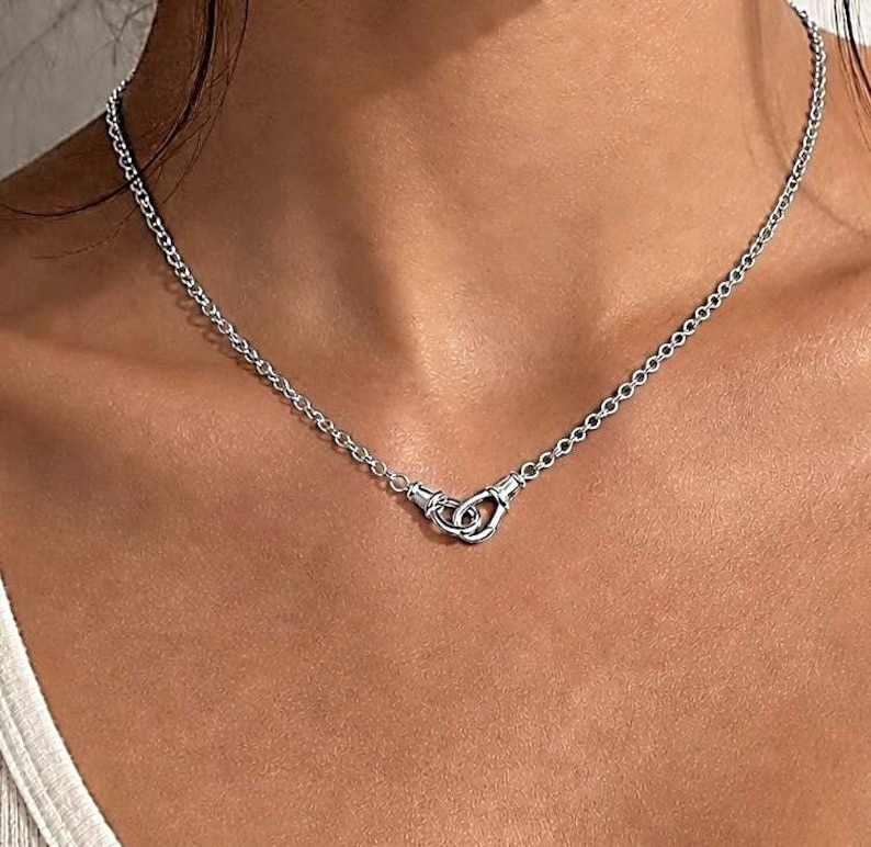 Puede incluir: Un collar plateado con una cadena delicada y un dise&ntilde;o de cierre &uacute;nico. El cierre presenta dos bucles entrelazados, creando una est&eacute;tica moderna y minimalista. El collar se lleva alrededor del cuello.