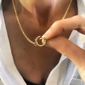 May include: A gold-toned necklace featuring a delicate chain and a circular clasp mechanism. The circular component is held between fingers, showcasing the polished metallic finish and the interlocking design of the chain links and the round closure.
