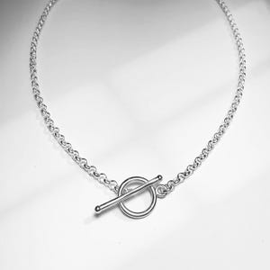 May include: A silver-toned chain necklace featuring a circular ring and a straight bar toggle clasp closure. The necklace is constructed from a series of interconnected round links that form a uniform chain pattern, finished with a polished metallic surface.