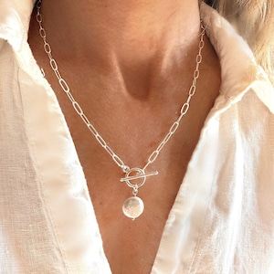 May include: A silver-toned necklace featuring a long oval link chain and a toggle clasp closure. A single, round, white, textured pearl-like pendant hangs from the centre of the toggle ring, resting against the collarbone area of the wearer.