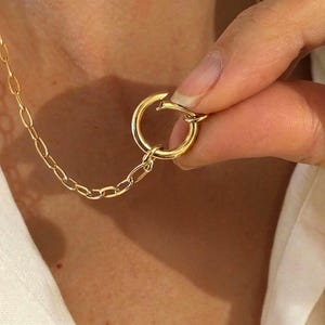 Peut inclure: Un collier doré avec un grand fermoir circulaire et une chaîne délicate. Le fermoir est ouvert, révélant le mécanisme de connexion. Le collier est présenté sur un fond blanc.