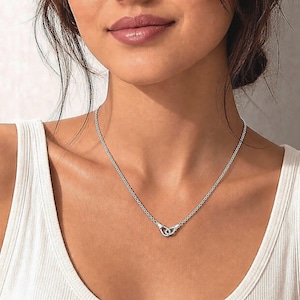 Puede incluir: Un collar plateado con una cadena delicada y un colgante con forma de dos c&iacute;rculos entrelazados. El collar se lleva alrededor del cuello, y el colgante est&aacute; centrado en el pecho. El fondo es de color neutro.